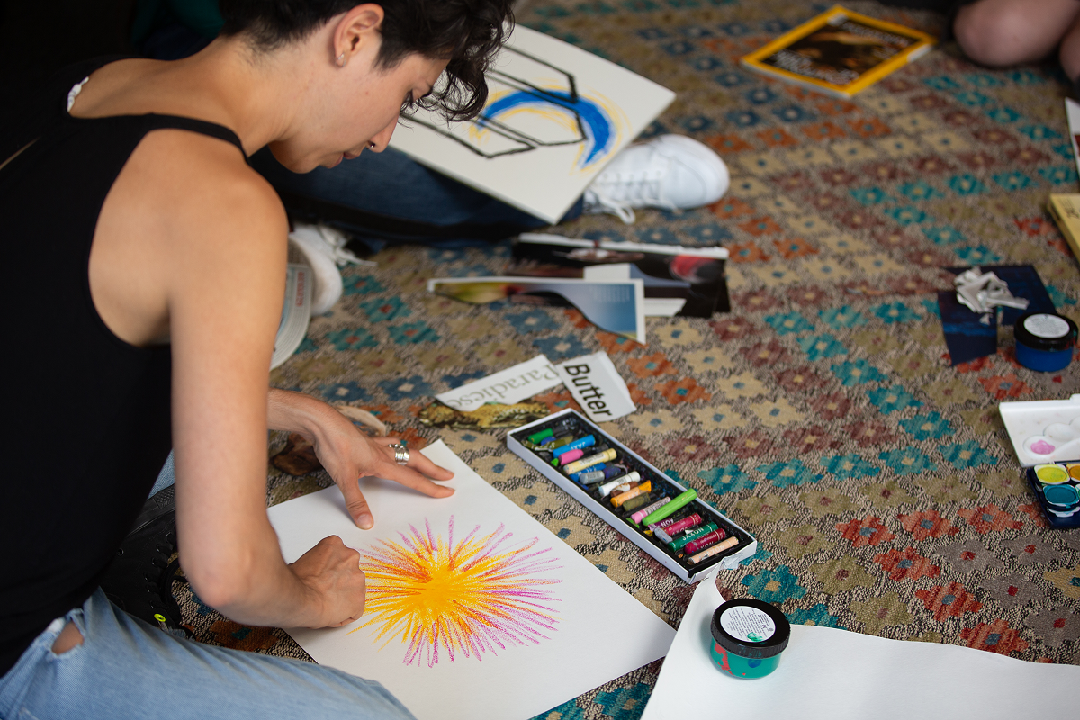 Eine Person sitzt auf einem gemusterten Teppich auf dem Boden und konzentriert sich während einer Kunsttherapiesitzung intensiv darauf, eine bunte Sonne mit Ölkreiden zu zeichnen.