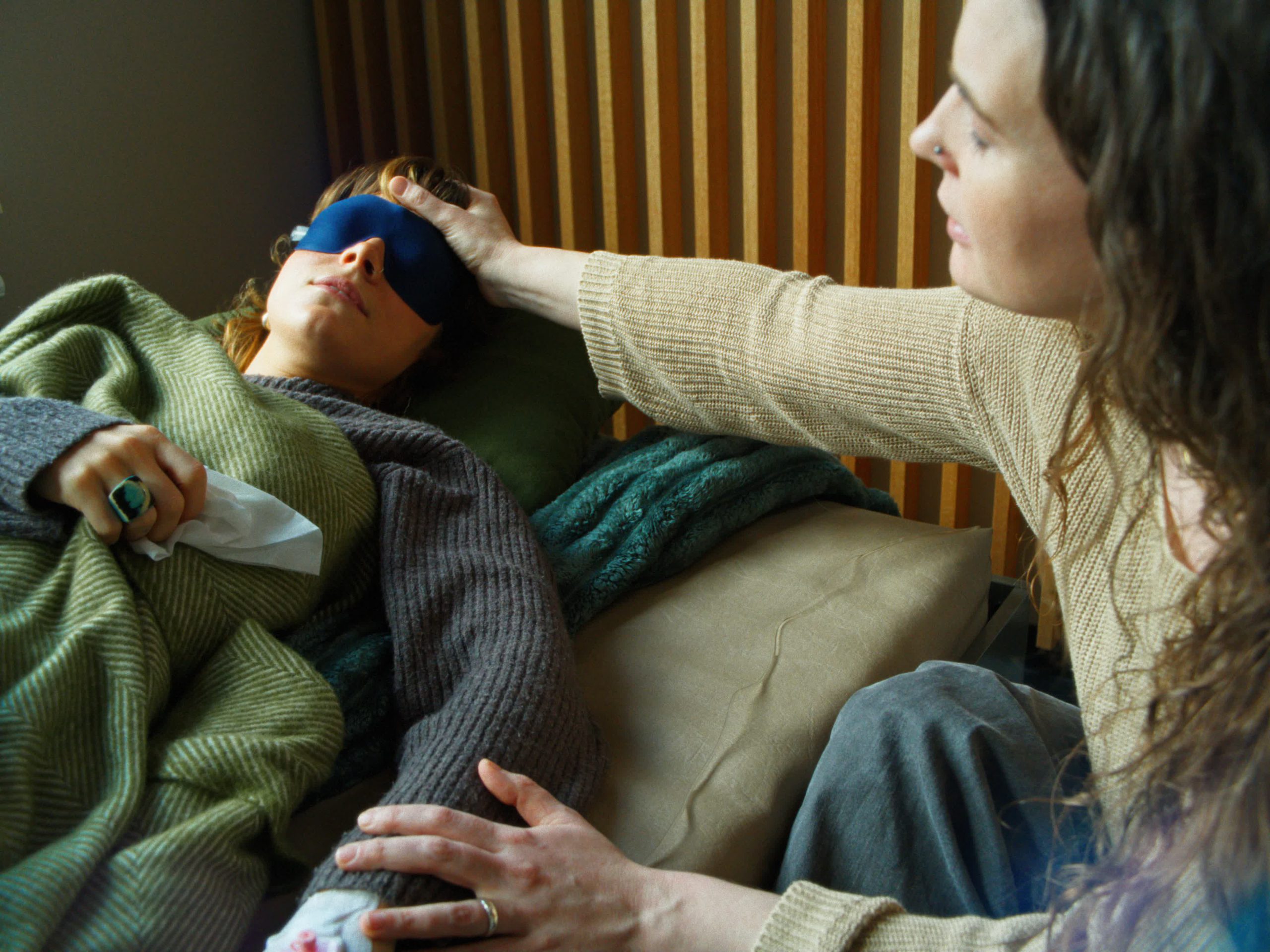 Eine Nahaufnahme, in der eine Therapeutin sanft ihre Hand auf die Stirn einer Patientin legt; die Patientin trägt eine blaue Schlafmaske und ruht unter einer grünen Decke.