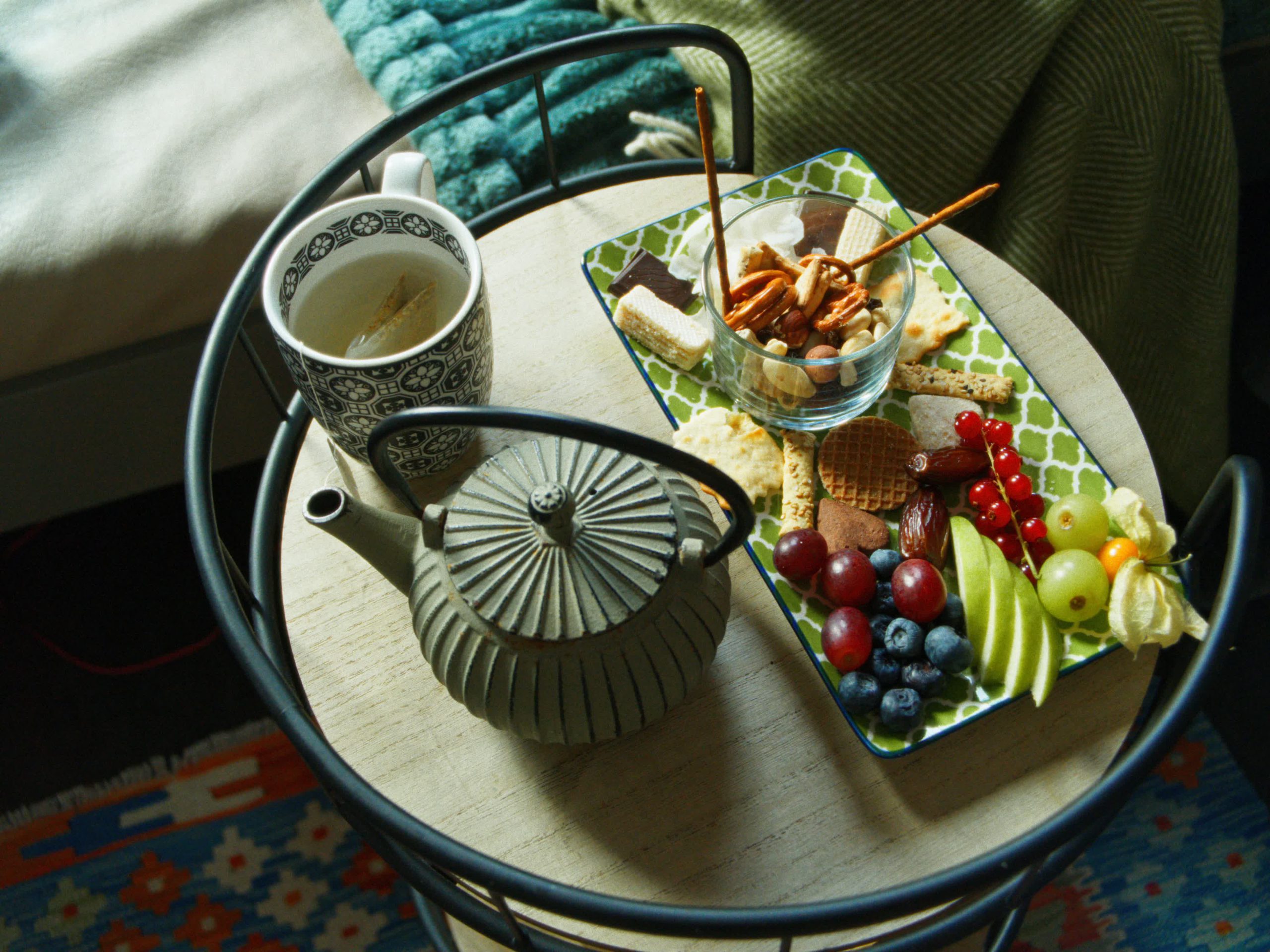 Eine Draufsicht auf ein Tablett mit einer dunklen Keramik-Teekanne, einer Tasse Tee und einer Schale mit gesunden Snacks wie Nüssen, Crackern und frischem Obst.