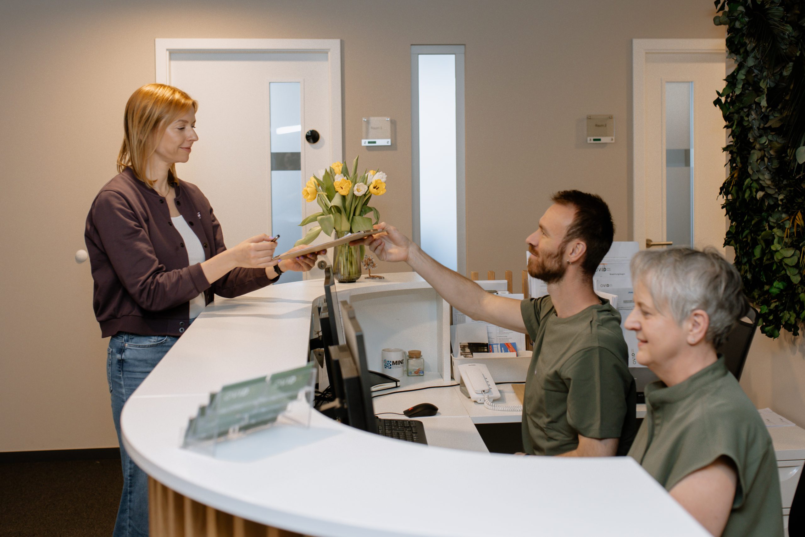 Ein Empfangsbereich, in dem eine Frau mit dem Personal spricht. Der Raum ist hell und professionell gestaltet und verfügt über einen geschwungenen Tresen sowie eine Vase mit Tulpen.