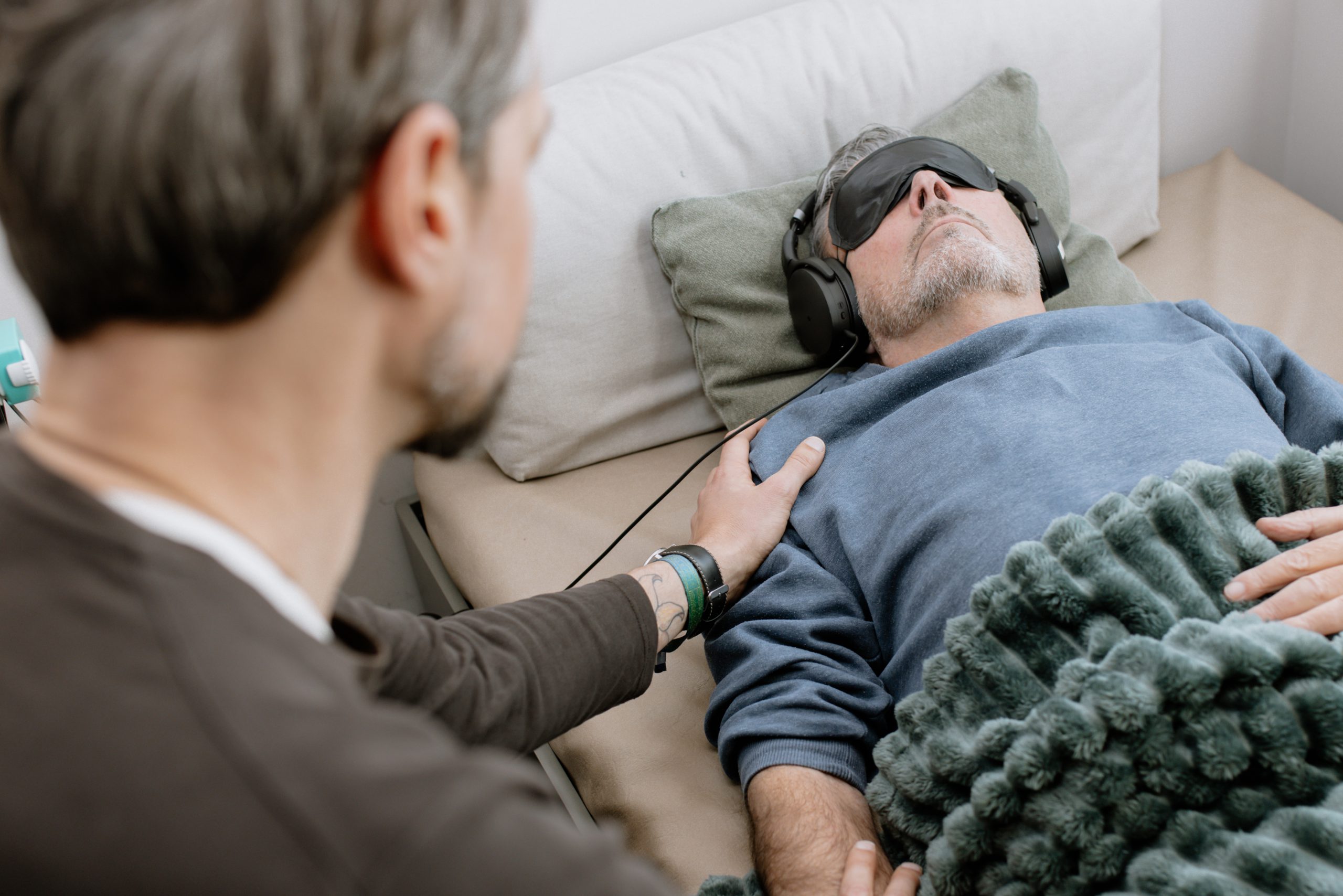 Ein Therapeut legt unterstützend seine Hand auf die Schulter eines Patienten, der mit Schlafmaske und Kopfhörern auf einer Behandlungsliege ruht.