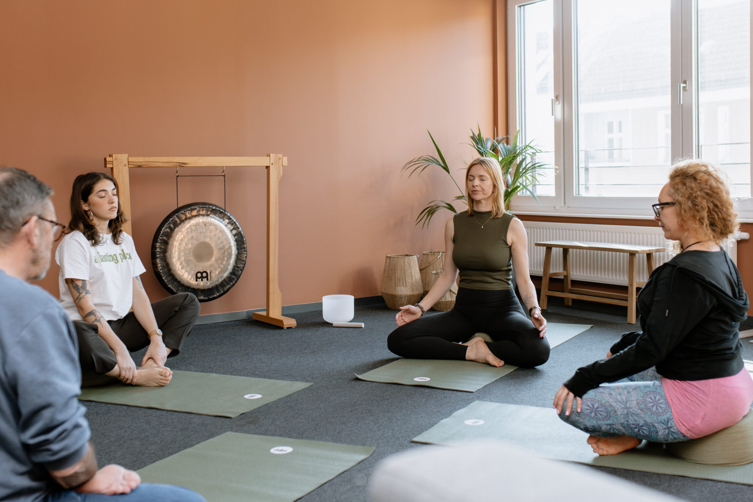 Eine Gruppe von Menschen sitzt auf Yogamatten in einem geräumigen, warm gestalteten Raum und nimmt an einer Gruppenmeditation oder Klangheilungssitzung teil, bei der im Hintergrund ein Gong zu sehen ist.