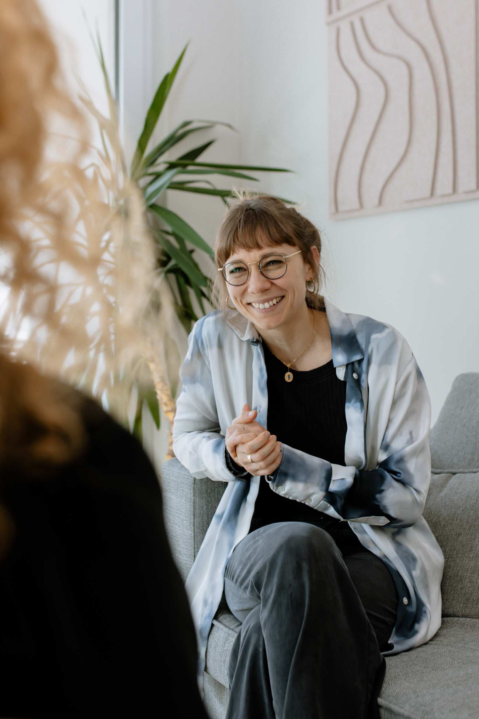 Eine lächelnde Frau mit Brille und Batikshirt sitzt bequem auf einem Sofa und schaut zu einer Person außerhalb des Bildausschnitts.