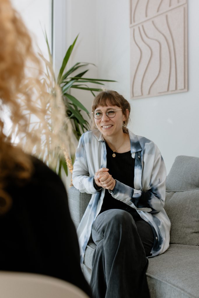 Eine lächelnde Frau mit Brille und Batikshirt sitzt bequem auf einem Sofa und schaut zu einer Person außerhalb des Bildausschnitts.