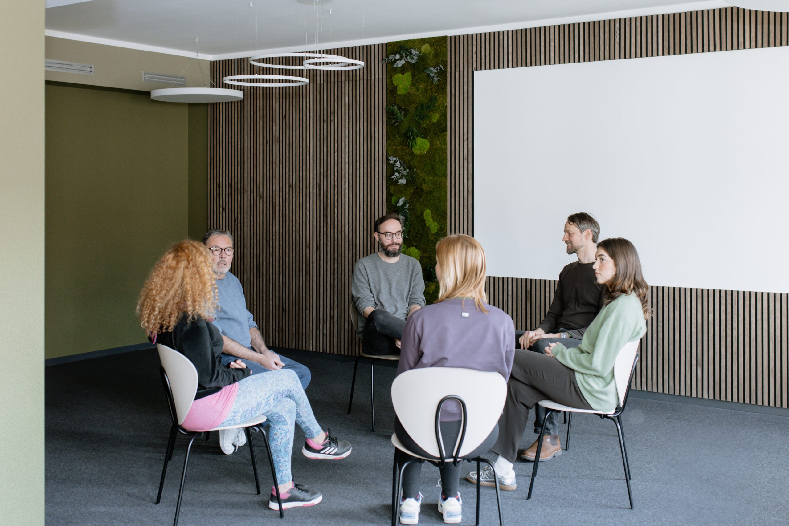 Eine Gruppe von Menschen sitzt im Kreis auf Stühlen in einem modernen Raum und nimmt an einer moderierten Gruppendiskussion oder Therapiesitzung teil.