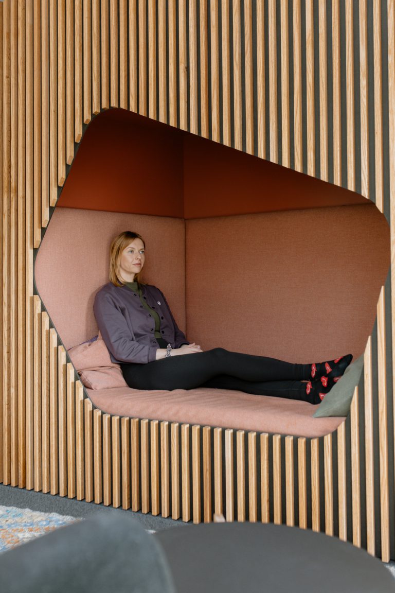 Eine Person sitzt ruhig in einer modernen, bequemen, pfirsichfarbenen Nische, die in eine Wand aus vertikalen Holzlamellen eingelassen ist.