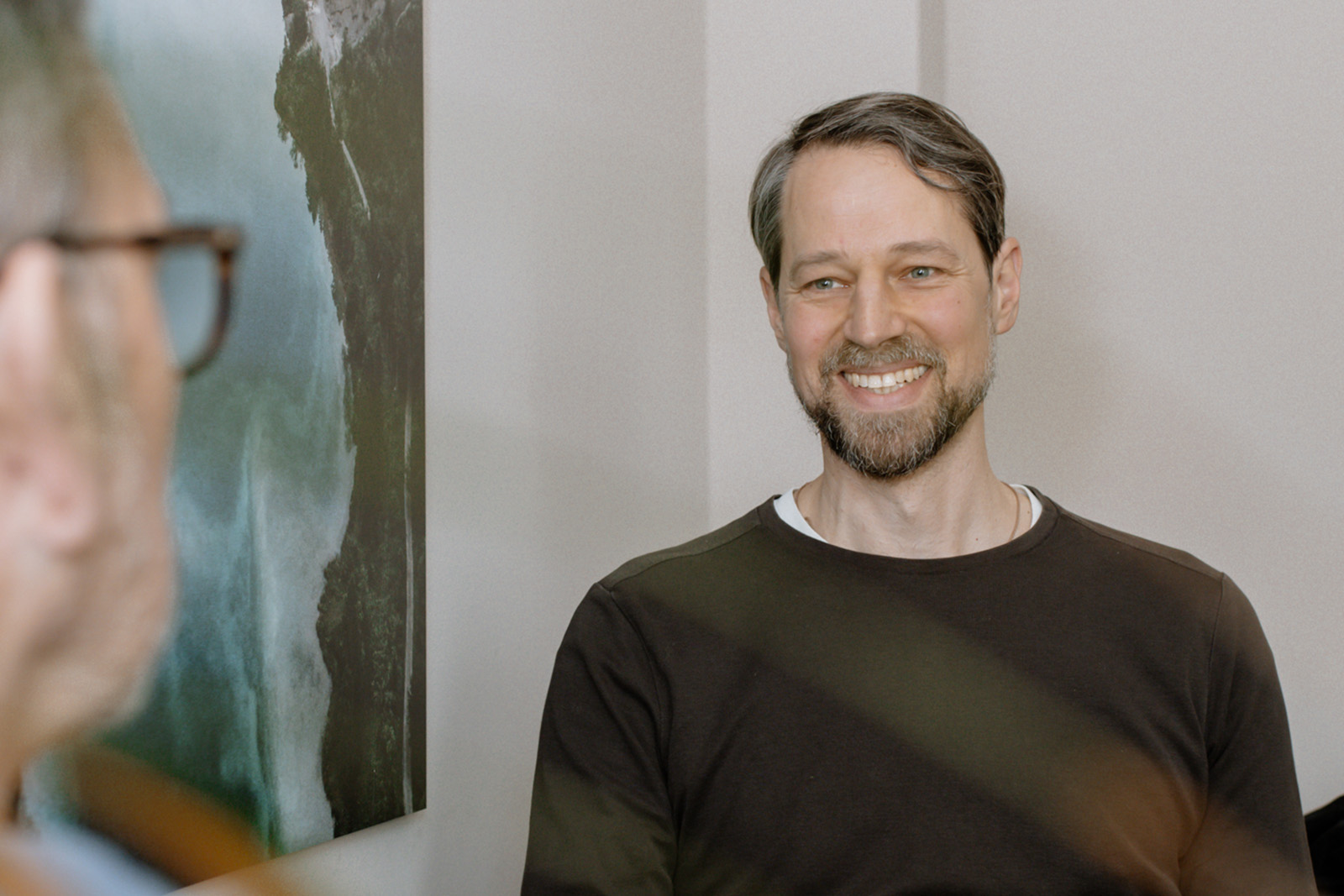 Ein bärtiger Mann lächelt herzlich während eines persönlichen Gesprächs mit einem Therapeuten (von hinten zu sehen).