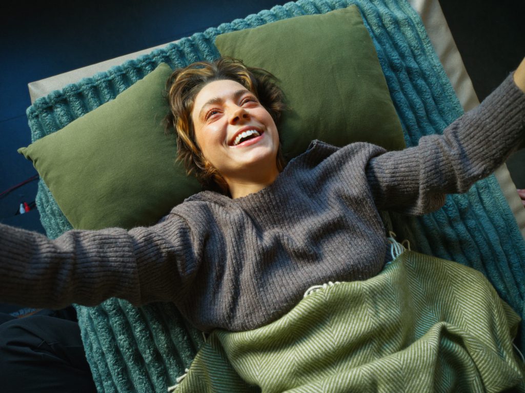 Eine Nahaufnahme aus der Vogelperspektive von einer jungen Person mit kurzen braunen Haaren, die lachend auf einer Behandlungsliege liegt. Sie ist unter einer grünen Decke mit Fischgrätmuster eingekuschelt, trägt einen grauen Rippstrickpullover und liegt auf zwei olivgrünen Kissen.